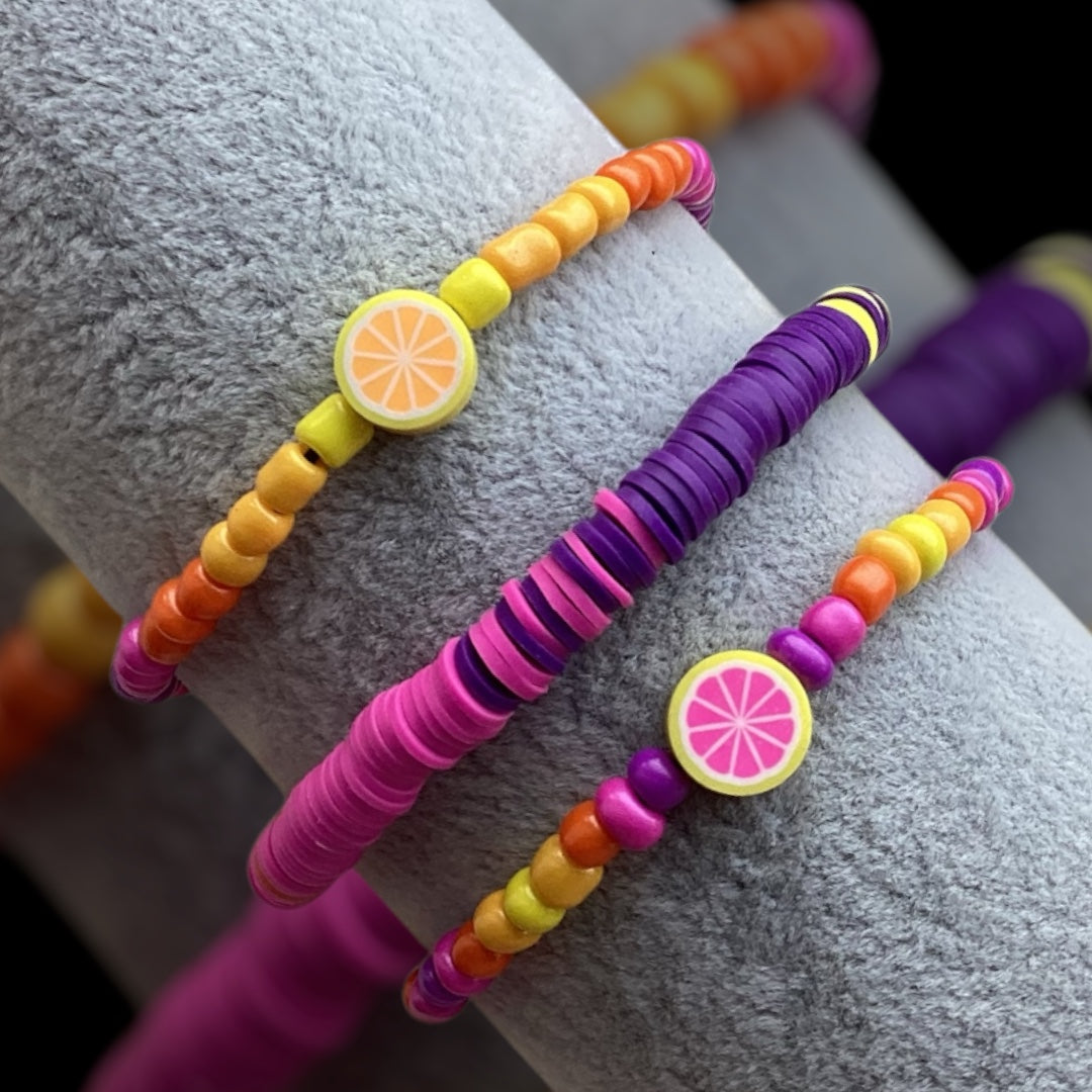 Set of three colorful handmade bracelets with fruit accents.
Sunset-inspired bracelet stack featuring yellow, orange, pink, and purple beads.
Handcrafted summer bracelet set with vibrant hues and playful designs.