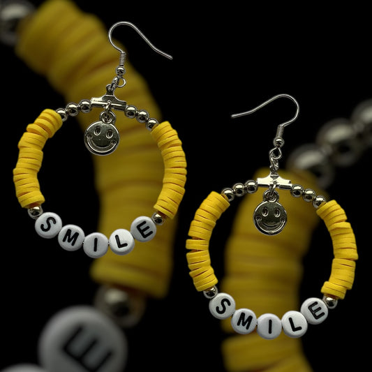 Handmade yellow hoop earrings with white “SMILE” beads and silver smiley face charm, made with polymer clay and silver-tone accents, cheerful and playful jewellery on a black background
